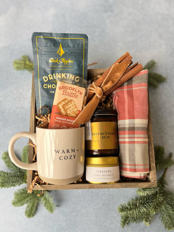 Cozy Winter Gift Box includes ceramic mug with "warm and cozy" printed on it, hot chocolate mix, buttered rum mix, brooklyn brittle cookies ginger spice cookies, one evergreen scented candle, and a few cinnamon sticks